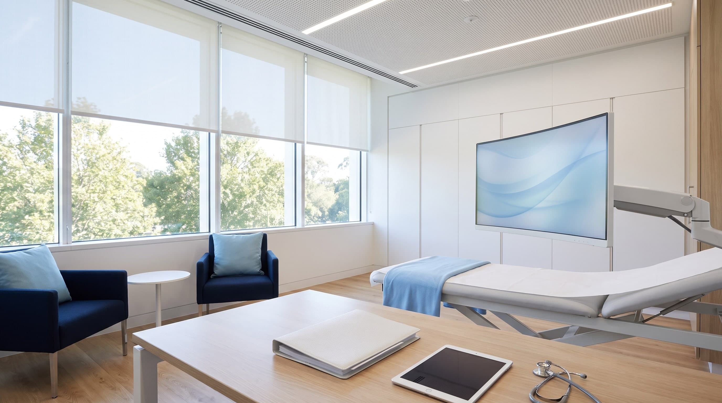 Großes, helles Untersuchungszimmer einer Klinik mit Liege, Bildschirm und Schreibtisch mit Stethoskop und Tablet, ohne Menschen